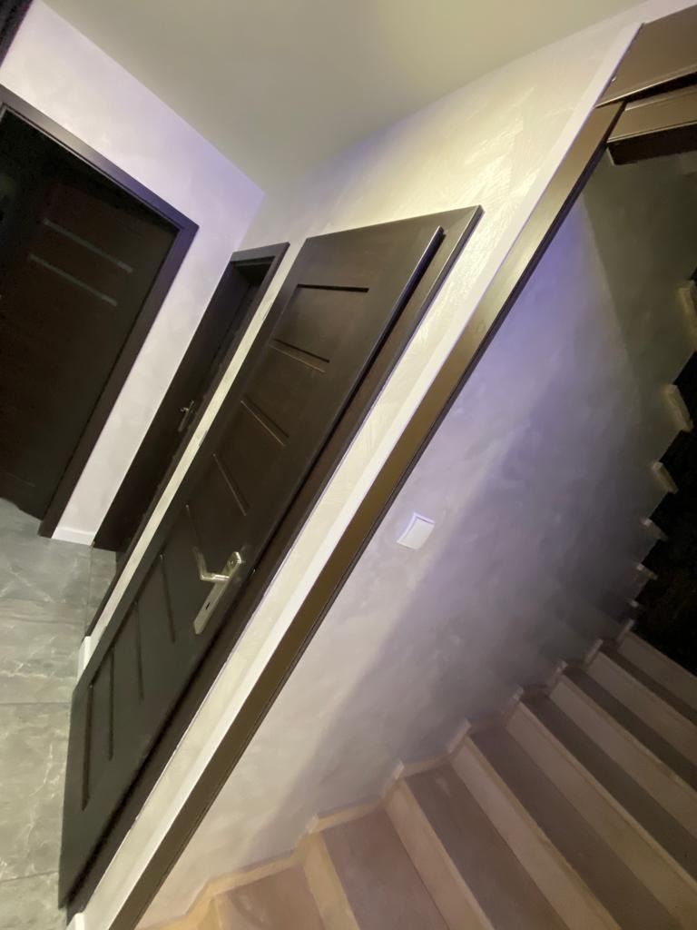 Ciemne, drewniane drzwi wewnętrzne z srebrną klamką, zamontowane w jasnym pomieszczeniu z widocznymi schodami i fakturą na ścianie.