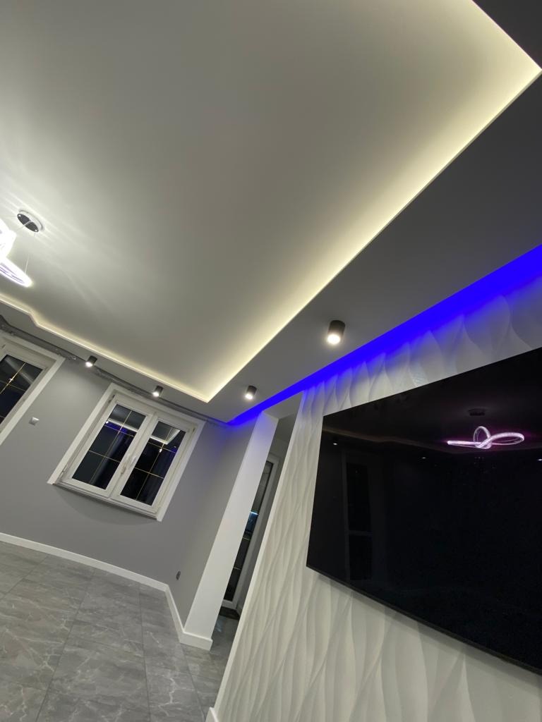 Nowoczesne wnętrze z podwieszanym sufitem z oświetleniem LED (białym i niebieskim), szarą ścianą, białymi oknami i telewizorem na ozdobnej ścianie.