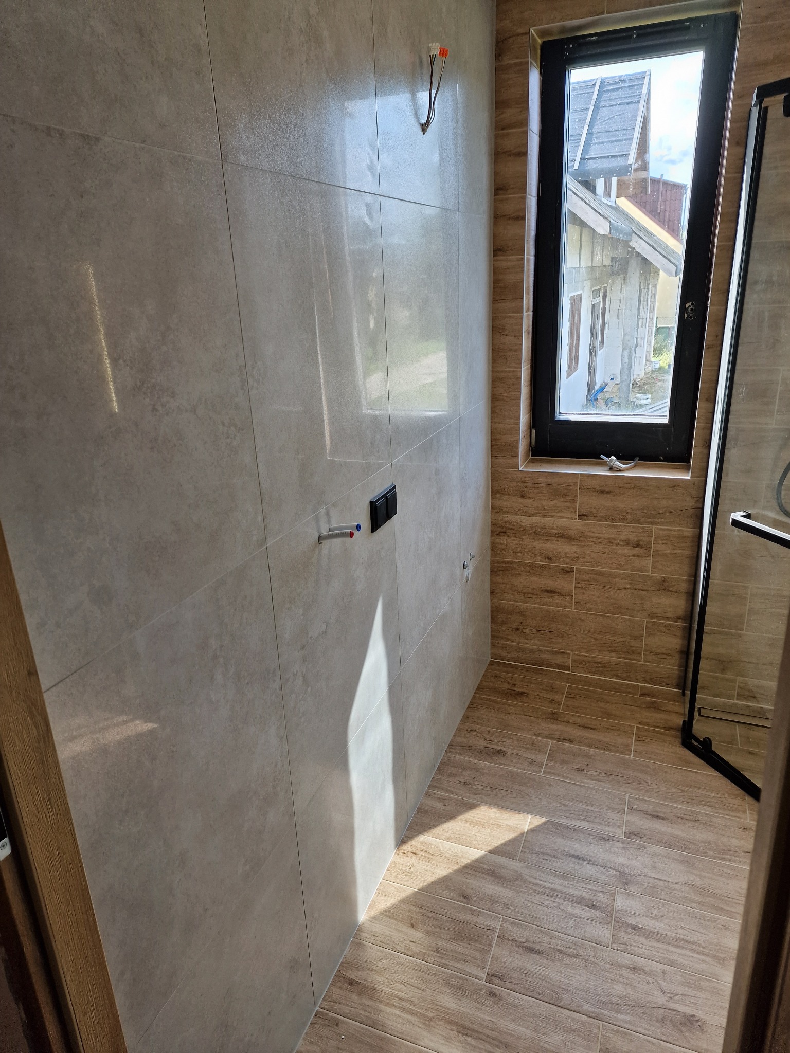 Wykończona łazienka z beżowymi płytkami imitującymi drewno na podłodze i ścianach, czarna rama okienna z widokiem na budynek z zewnątrz, przygotowane instalacje pod armaturę.