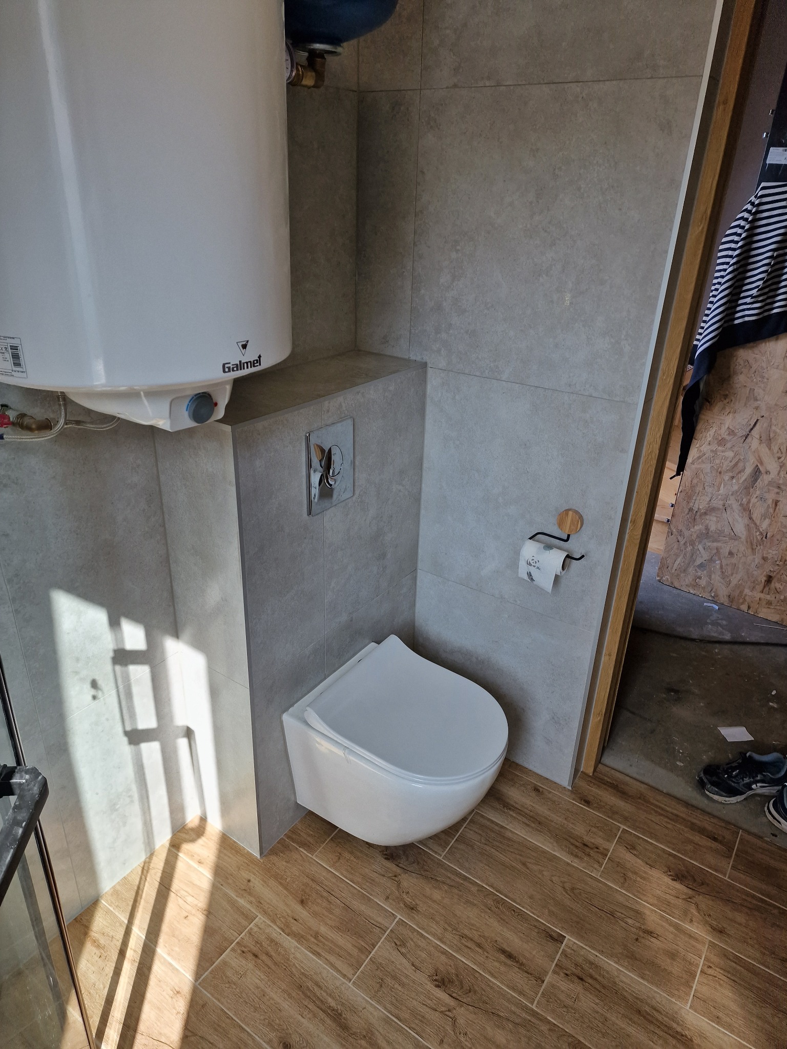 Nowoczesna, minimalistyczna toaleta podwieszana w szarej łazience z drewnopodobną podłogą, widoczny bojler Galmet i cień na ścianie.