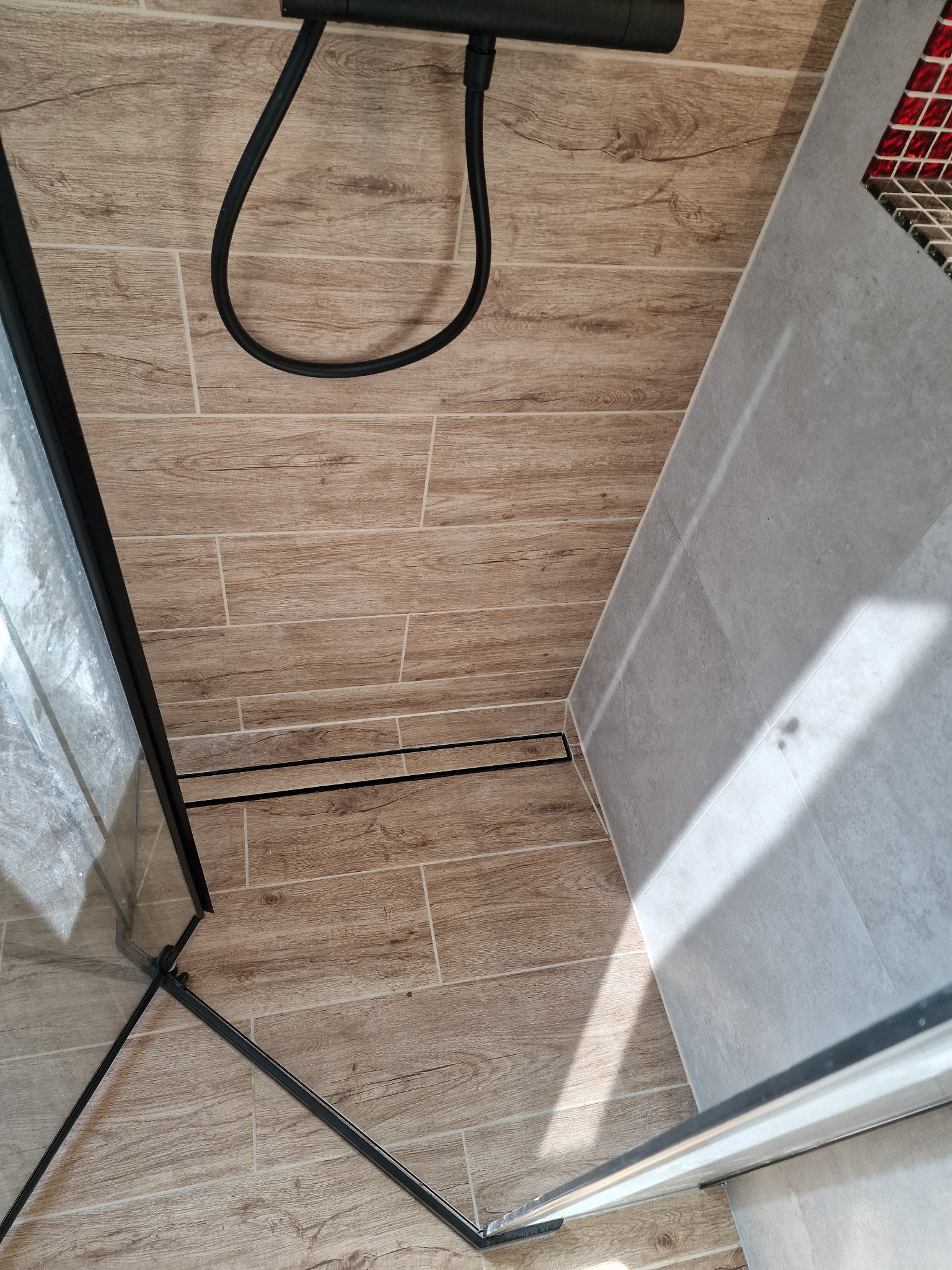 Kabina prysznicowa typu walk-in z czarnymi profilami i odpływem liniowym w podłodze wyłożonej płytkami imitującymi drewno, ściana wykończona szarymi płytkami.