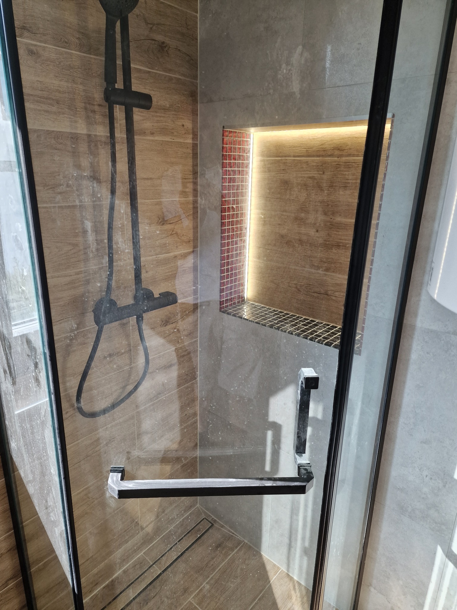 Kabina prysznicowa z czarną armaturą, drewnopodobnymi płytkami i wnęką z podświetleniem LED wykończoną mozaiką.