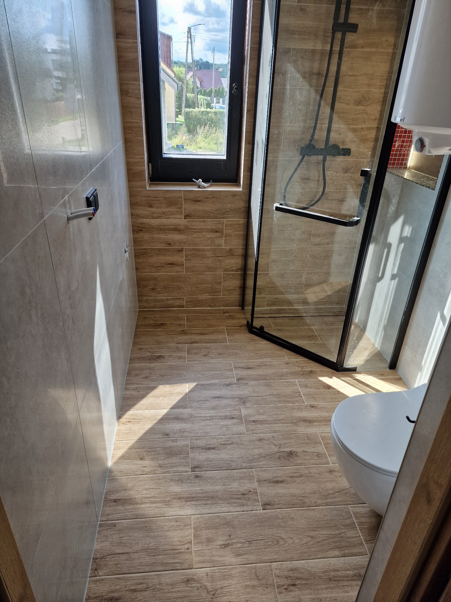 Wykończona łazienka z kabiną prysznicową typu walk-in, płytkami imitującymi drewno na podłodze i ścianach, oraz oknem z widokiem na zieleń.