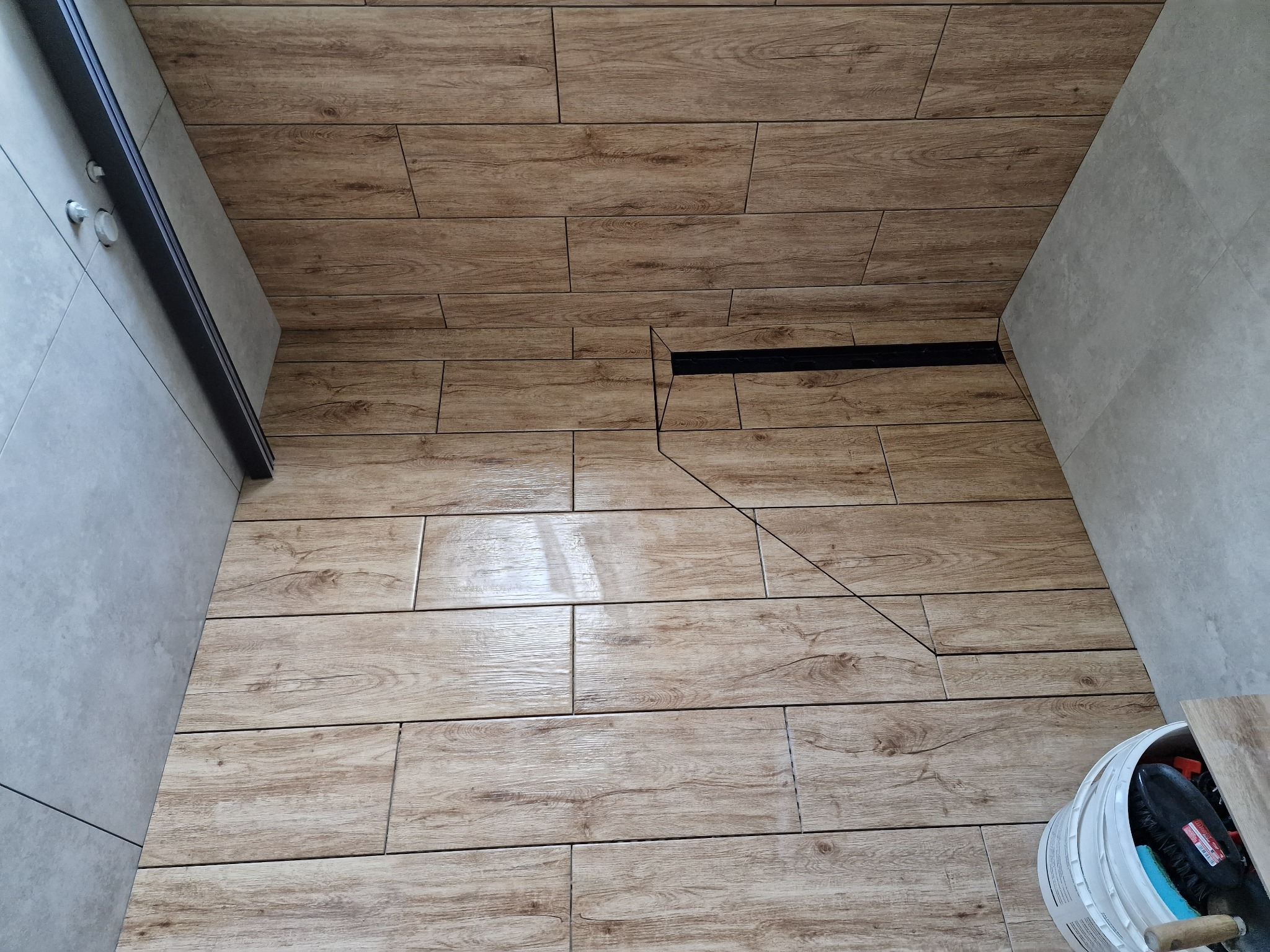 Ułożona pod kątem brązowa podłoga z płytek imitujących drewno w kabinie prysznicowej, widoczna czarna listwa odpływowa i wiadro z akcesoriami do fugowania.