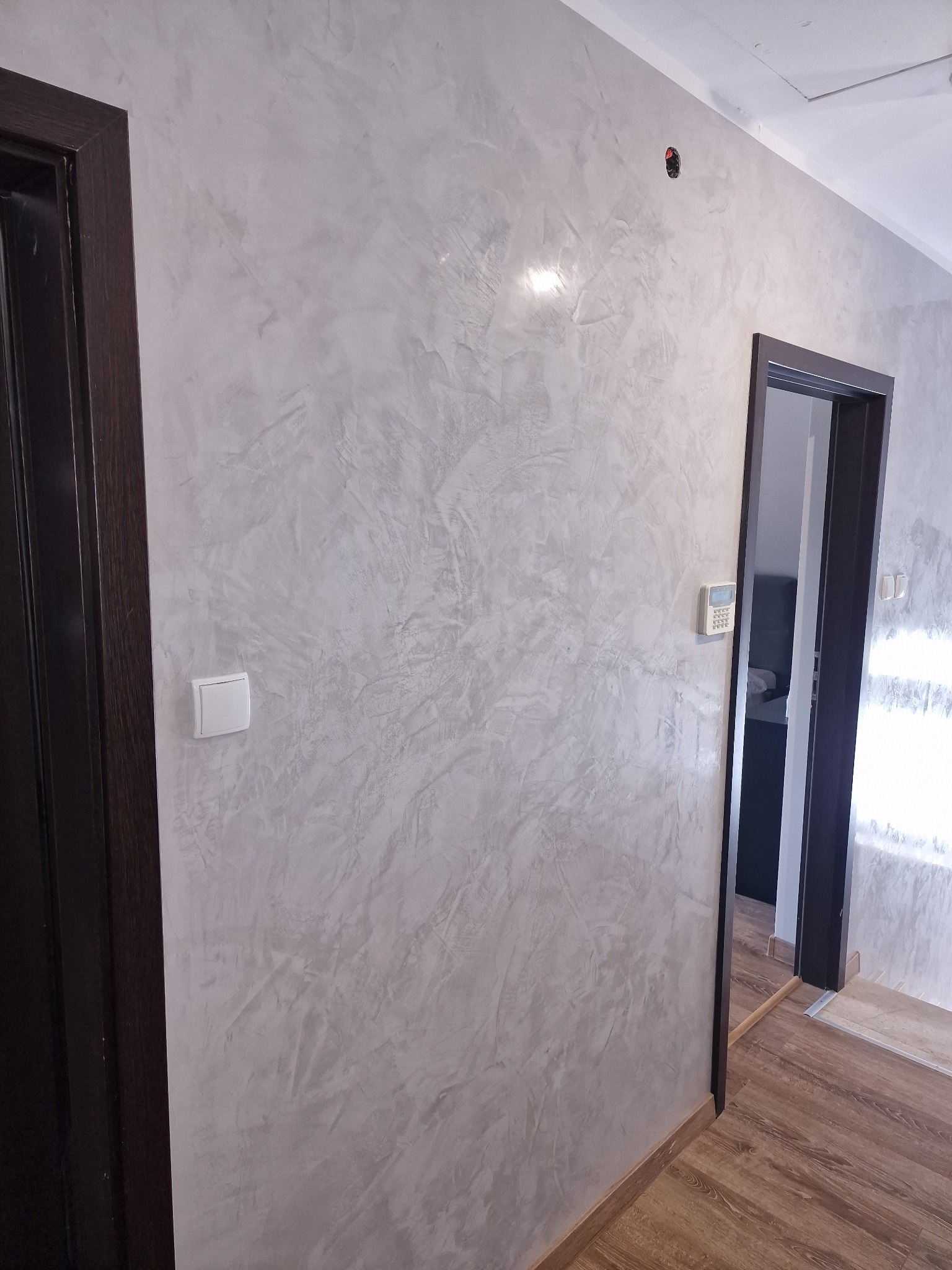 Ściana w korytarzu z dekoracyjną, szarą strukturą, widoczny włącznik światła, czarna futryna drzwi wejściowych i futryna drzwi do pomieszczenia z widocznym panelem sterowania.