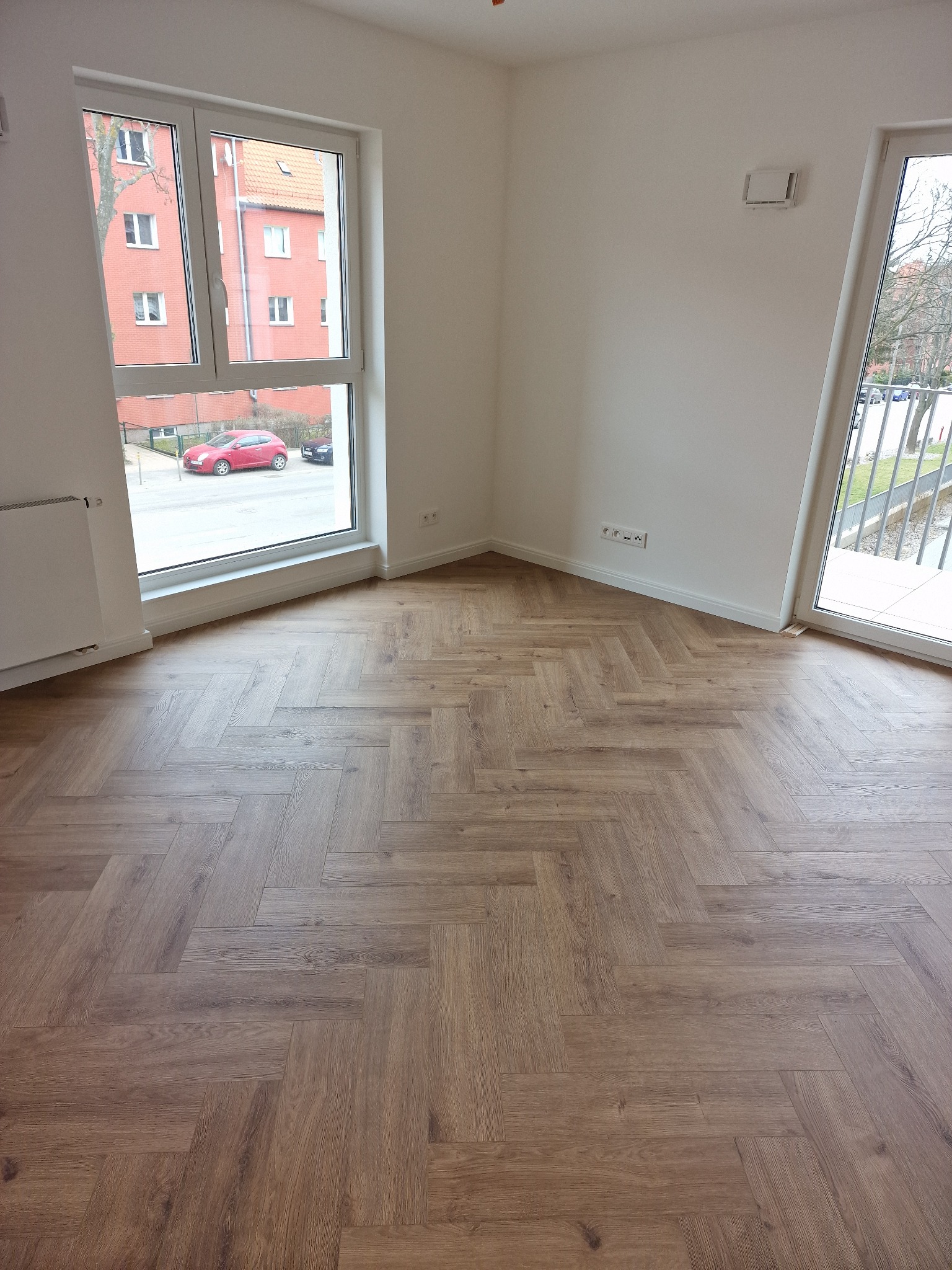 Pusty pokój z nową, ułożoną w jodełkę, drewnianą podłogą, białymi ścianami, dużym oknem z widokiem na ulicę i balkonem.