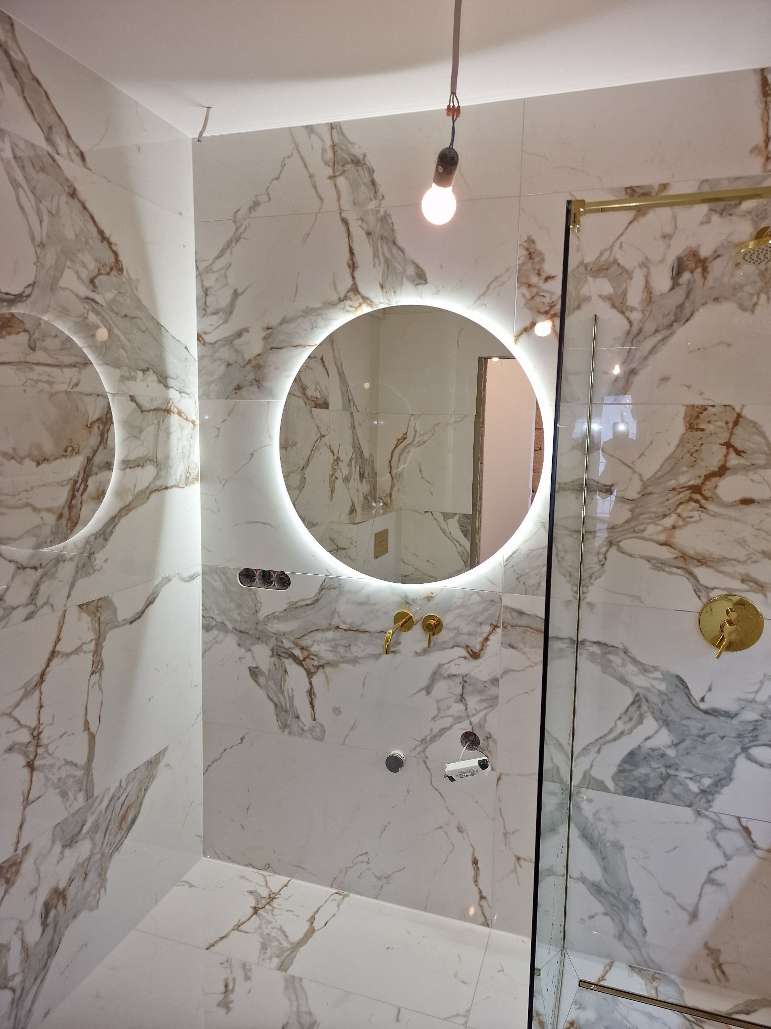 Nowoczesna łazienka z białymi płytkami imitującymi marmur, okrągłe lustro z podświetleniem LED, złota armatura i kabina prysznicowa ze złotymi profilami, surowa żarówka zwisająca z sufitu.