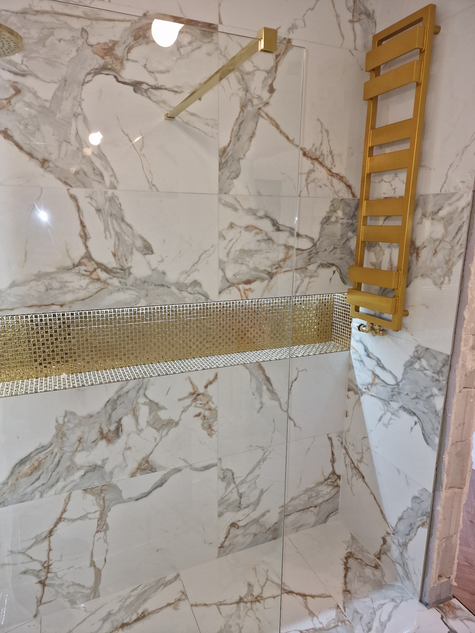 Fragment łazienki z marmurowymi płytkami, złotą mozaiką w formie półki i złotym grzejnikiem drabinkowym.