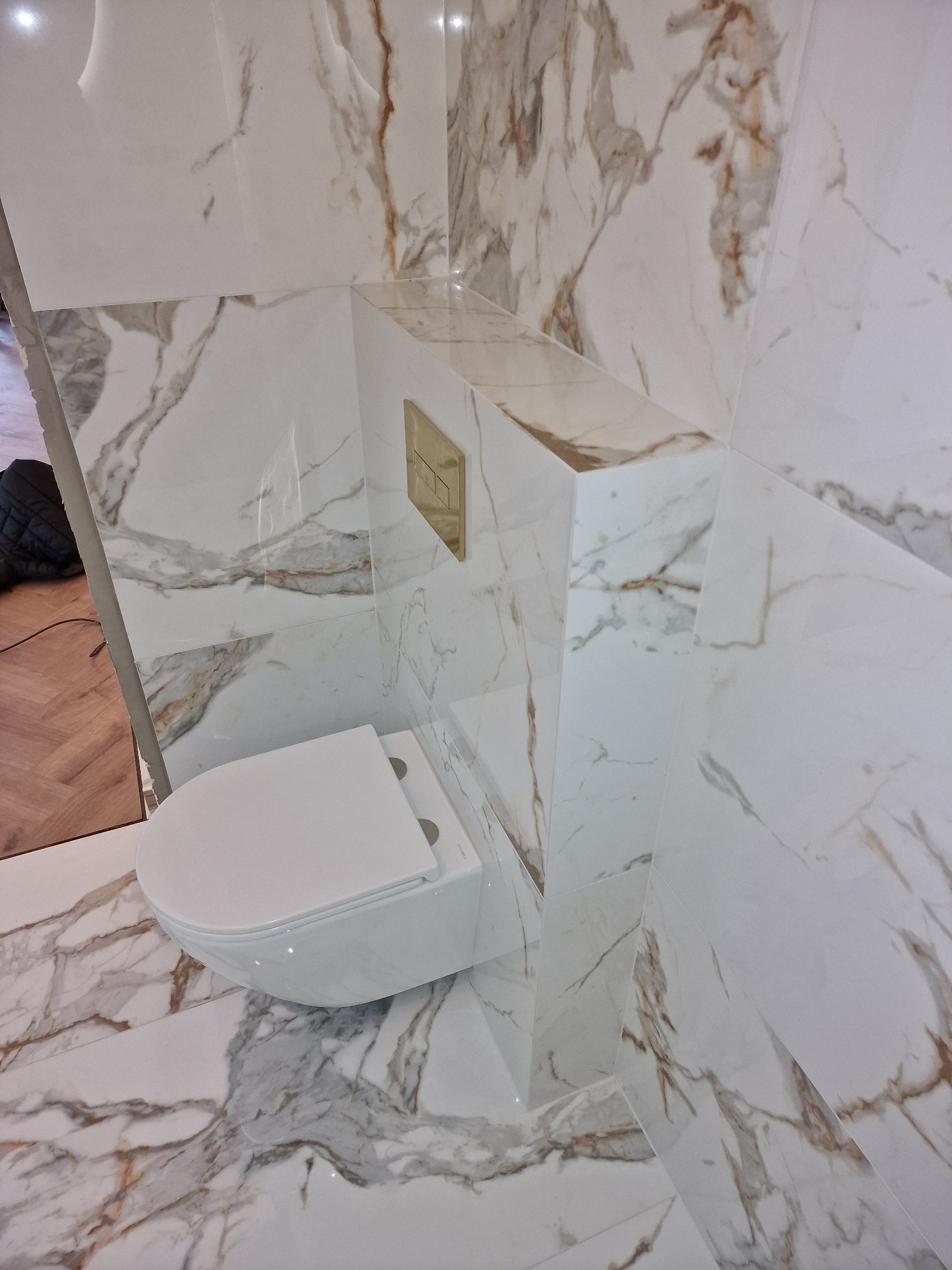 Podwieszana toaleta z białej ceramiki, zamontowana w rogu łazienki wyłożonej białymi płytkami imitującymi marmur z brązowymi żyłami, widoczny złoty przycisk spłuczki.