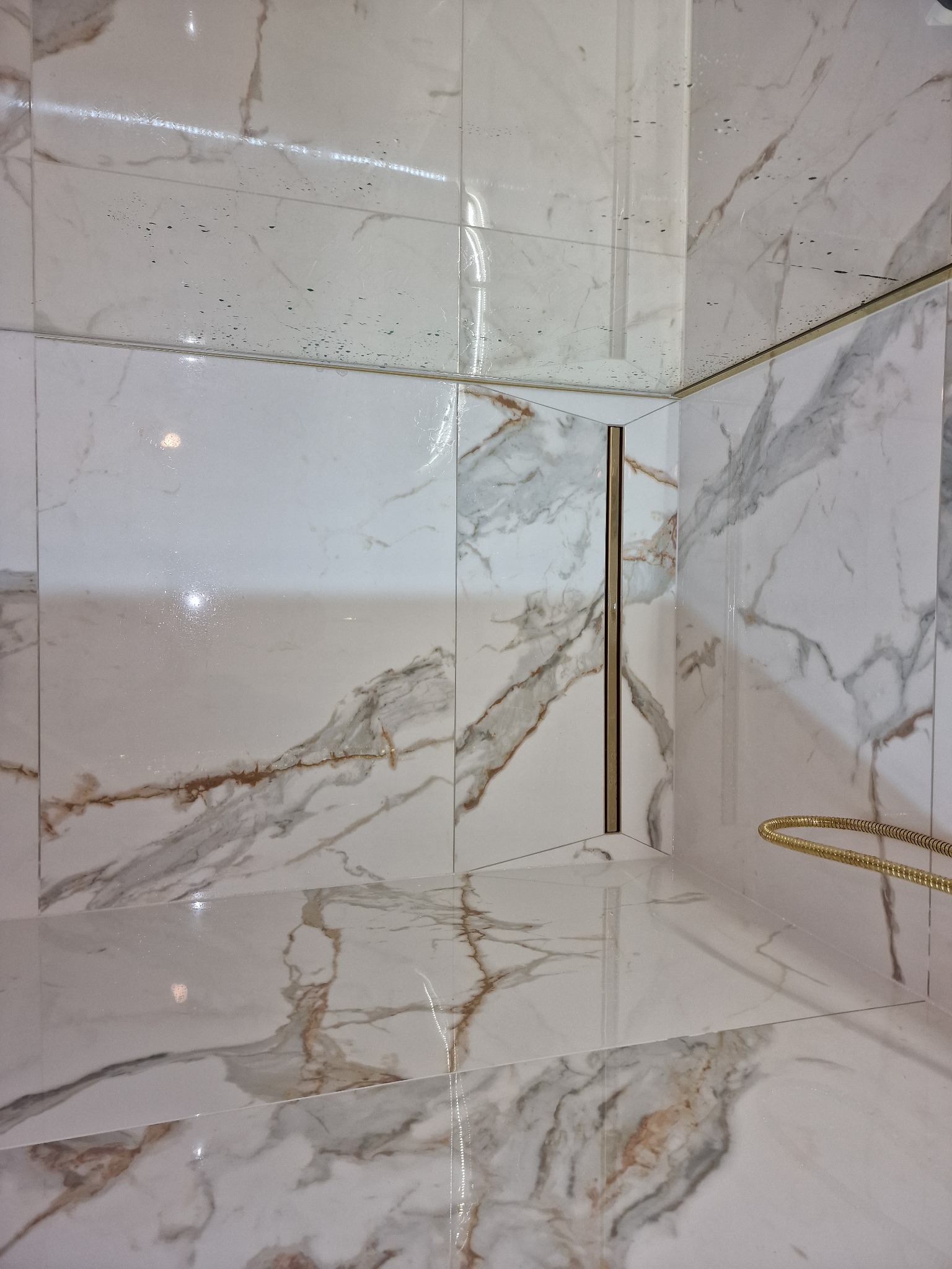 Wykończona kabina prysznicowa z białymi płytkami imitującymi marmur, złotym odpływem liniowym i słuchawką prysznicową, widoczne krople wody na szkle.