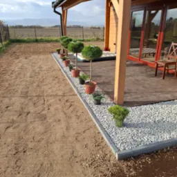 Świeżo posadzone drzewka w donicach wzdłuż obrzeża z kostki i białego grysu przy drewnianym tarasie, przygotowany teren pod trawnik w tle.