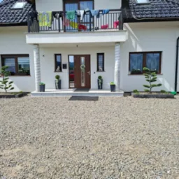 Elewacja domu z ozdobnymi drzewkami bonsai w donicach i kamiennym podłożem. Na balkonie suszą się ubrania. Widoczne góry w tle.