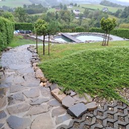 OZBUD Artur Bołoz - Ogród z oczkiem wodnym i kamienną ścieżką. Widok na wzgórza w tle. Elementy dekoracyjne z kamienia i roślinności. Ujęcie z góry, podkreślające kompozycję ogrodu.