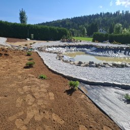 OZBUD Artur Bołoz - Etap budowy oczka wodnego z folią, kamieniami i ścieżką z grysu na tle zieleni. Widoczne przygotowane podłoże i małe sadzonki. Słoneczny dzień.