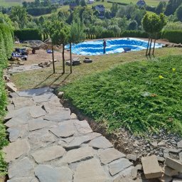 OZBUD Artur Bołoz - Budowa basenu w ogrodzie z widokiem na pagórkowaty krajobraz. Wyłożona kamienna ścieżka prowadzi do niecki basenowej wyłożonej niebieską folią.