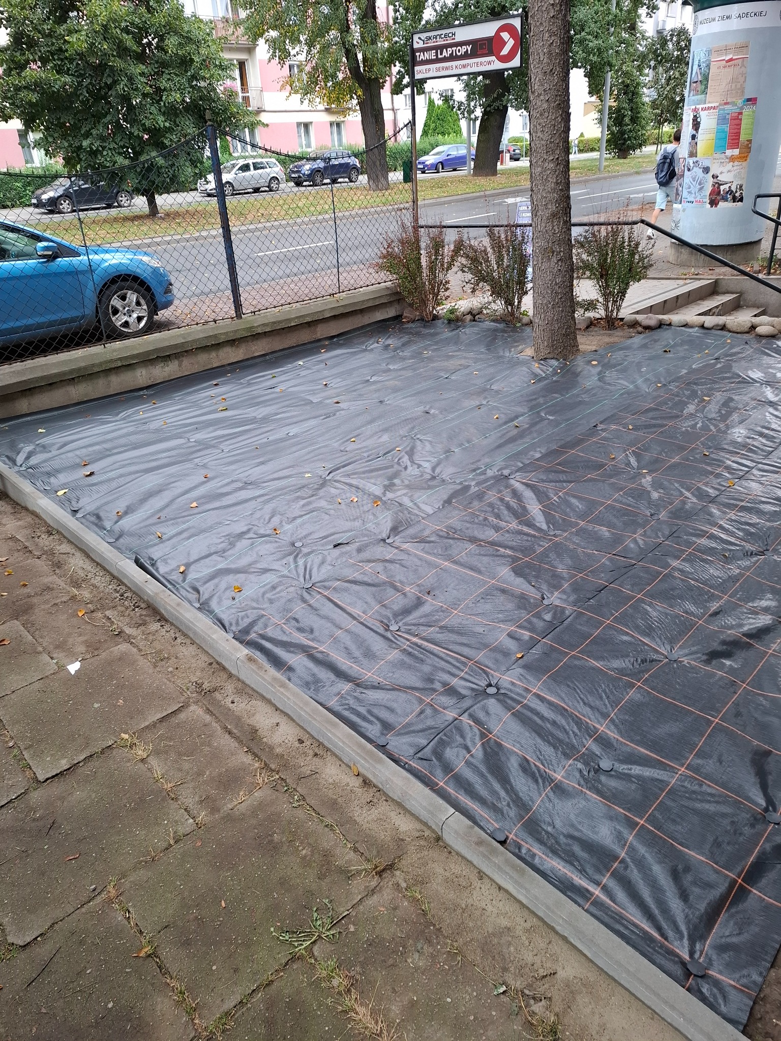 Przygotowanie terenu pod ogród: czarna agrowłóknina z pomarańczową siatką, przymocowana do podłoża szpilkami, otoczona betonowym obrzeżem, z drzewem rosnącym pośrodku.