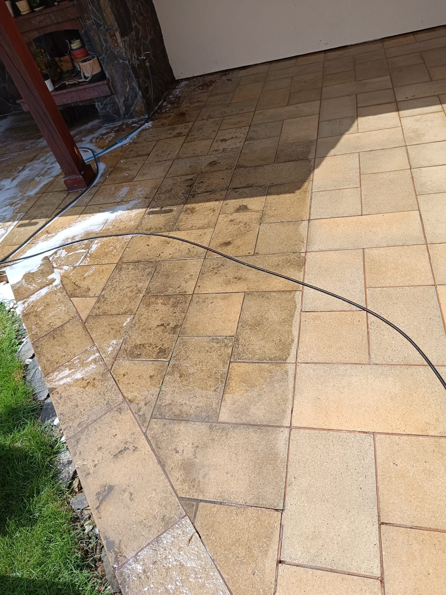 Czyszczenie kostki brukowej na tarasie za pomocą myjki ciśnieniowej, widoczny efekt przed i po czyszczeniu, z pianą i wężem. Część kostki po lewej stronie jest mokra i czysta, a po prawej sucha...
