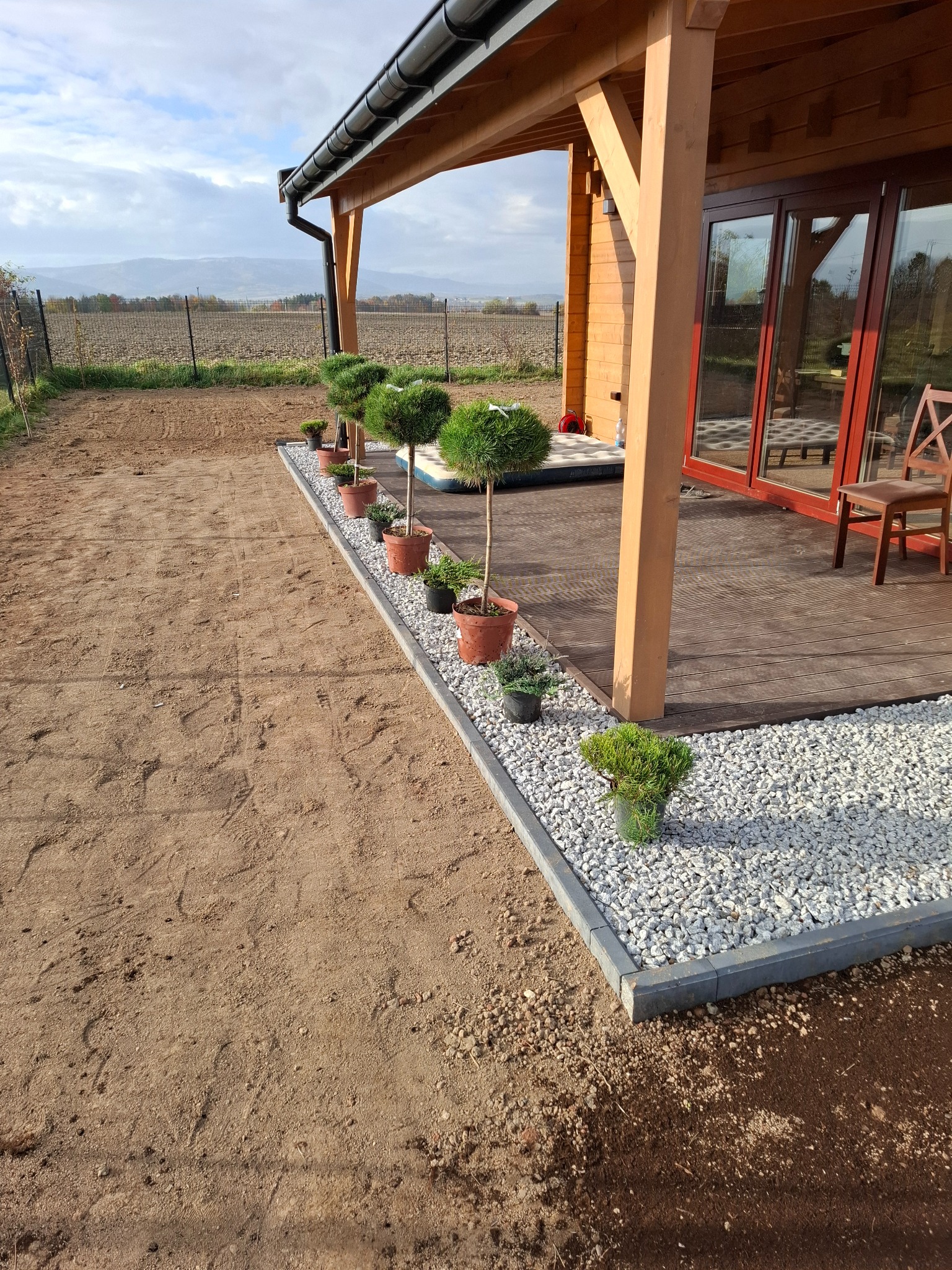 Świeżo posadzone drzewka w donicach wzdłuż obrzeża z kostki i białego grysu przy drewnianym tarasie, przygotowany teren pod trawnik w tle.