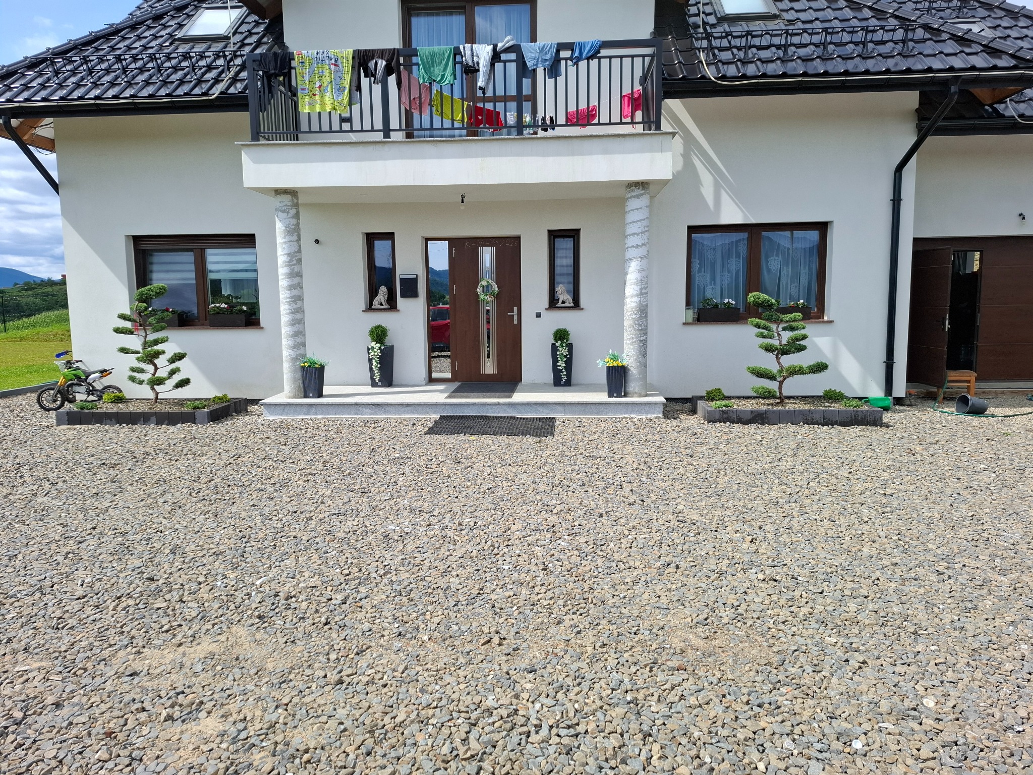 Elewacja domu z ozdobnymi drzewkami bonsai w donicach i kamiennym podłożem. Na balkonie suszą się ubrania. Widoczne góry w tle.