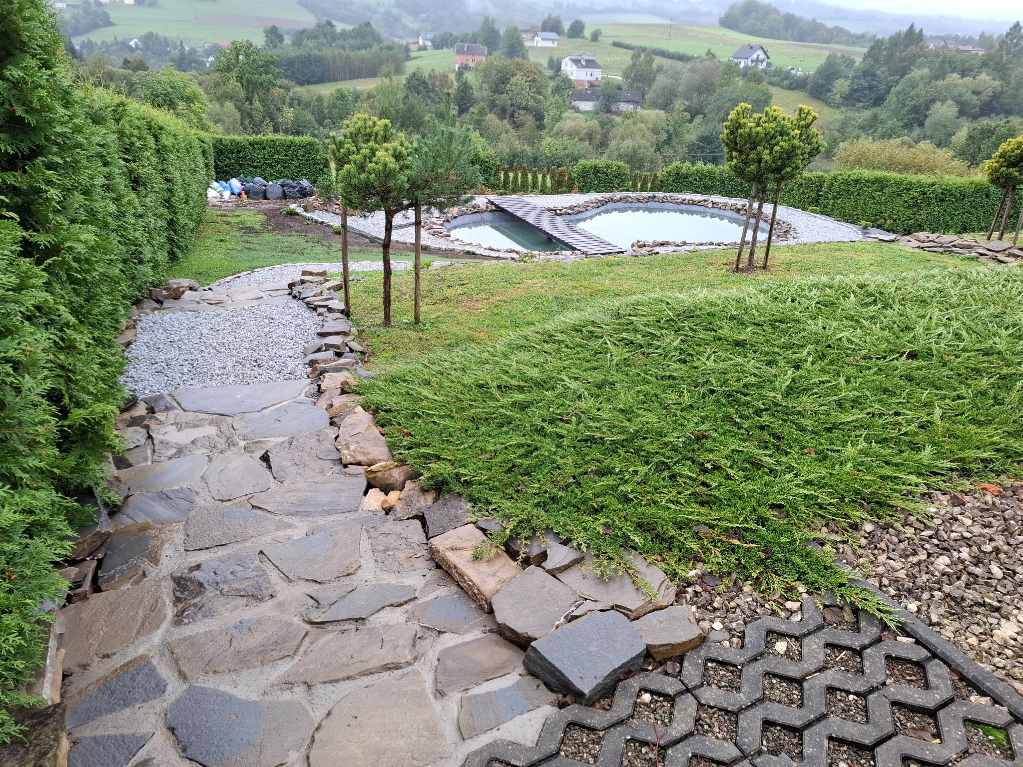 Ogród z oczkiem wodnym i kamienną ścieżką. Widok na wzgórza w tle. Elementy dekoracyjne z kamienia i roślinności. Ujęcie z góry, podkreślające kompozycję ogrodu.
