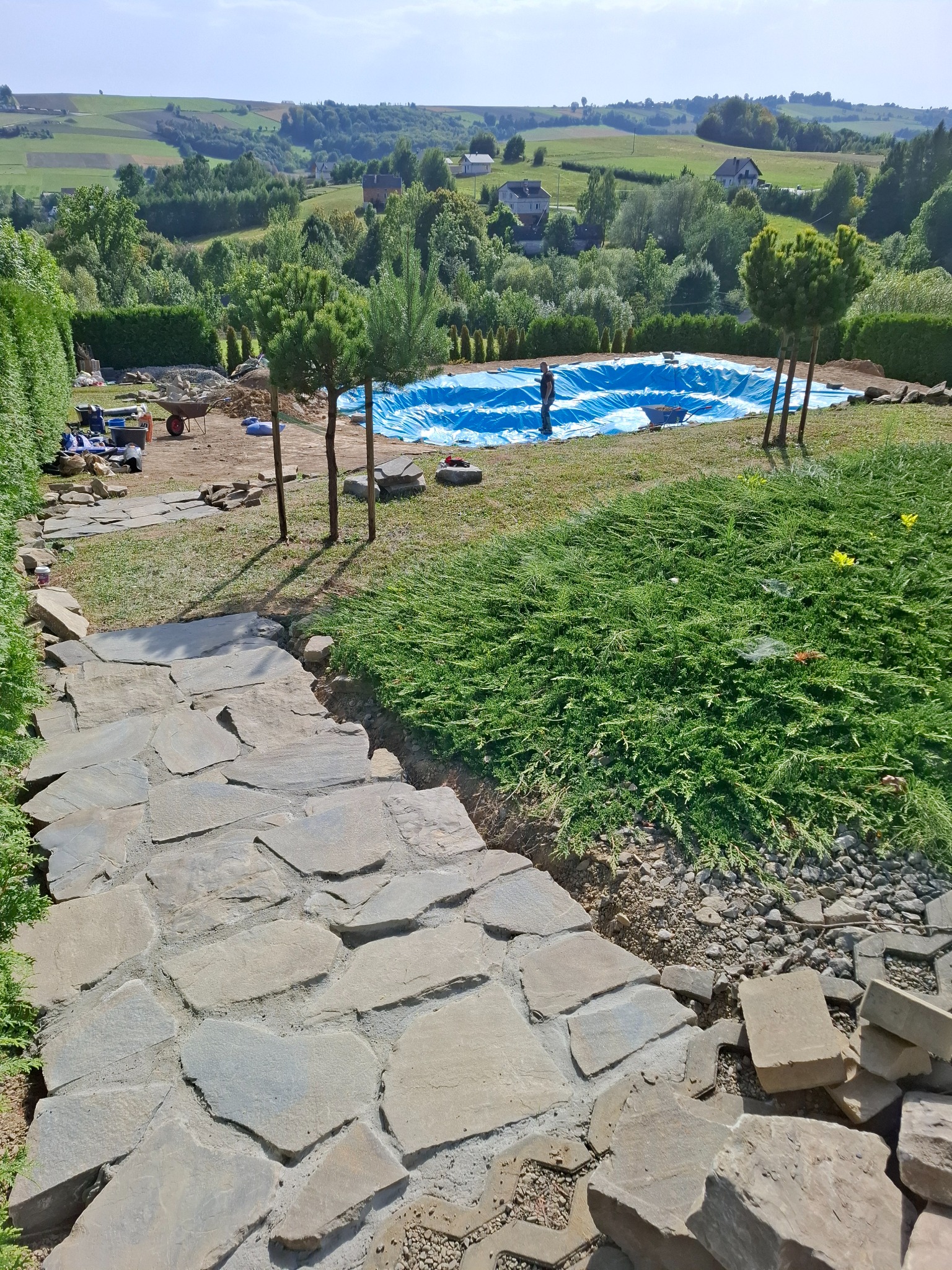 Budowa basenu w ogrodzie z widokiem na pagórkowaty krajobraz. Wyłożona kamienna ścieżka prowadzi do niecki basenowej wyłożonej niebieską folią.