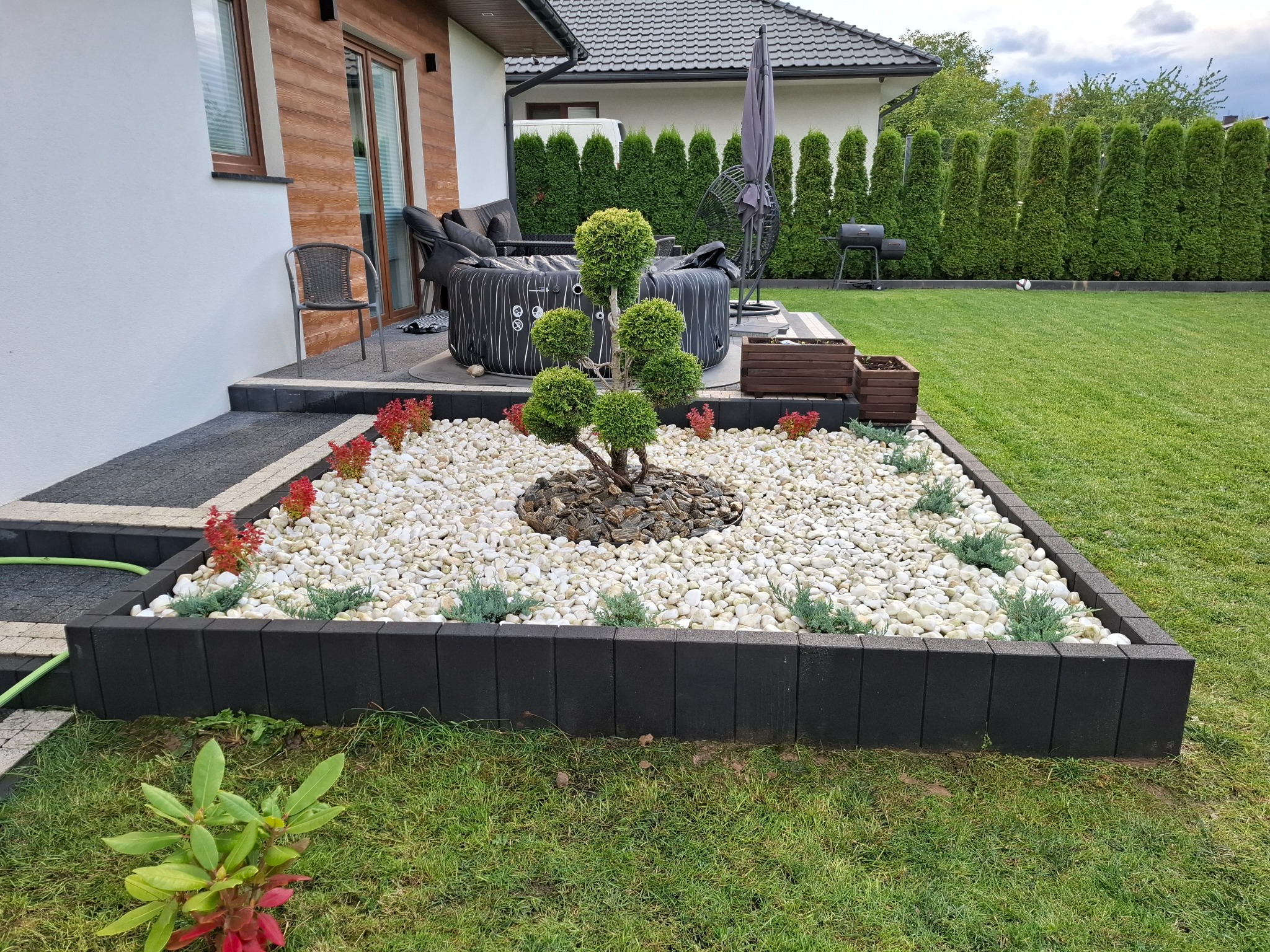 Ogród z ozdobnym drzewkiem formowanym, białymi kamieniami i obrzeżem z czarnej kostki. W tle trawnik, tuje i jacuzzi.