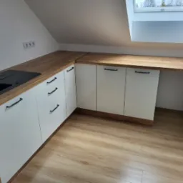 Nowoczesna zabudowa kuchenna na poddaszu ze skośnym sufitem i oknem dachowym, fronty szafek białe, blat drewnopodobny, czarny zlew.