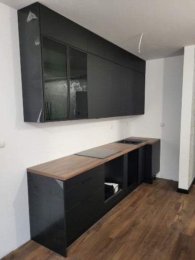 Częściowo zmontowana, nowoczesna, czarna kuchnia z drewnianym blatem, widoczne folie ochronne na górnych szafkach, surowy kabel zwisający z sufitu.