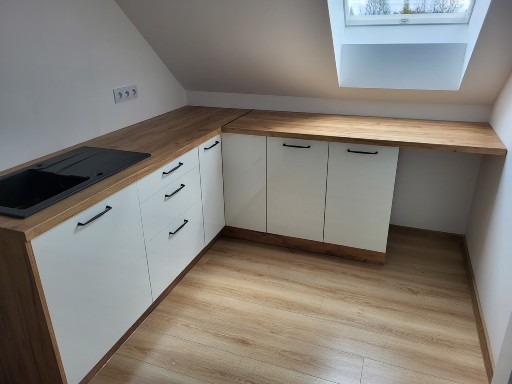 Nowoczesna zabudowa kuchenna na poddaszu ze skośnym sufitem i oknem dachowym, fronty szafek białe, blat drewnopodobny, czarny zlew.