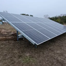 Panel słoneczny na metalowej konstrukcji, zamontowany na gruncie, w tle budynek mieszkalny, traktor i drzewa.