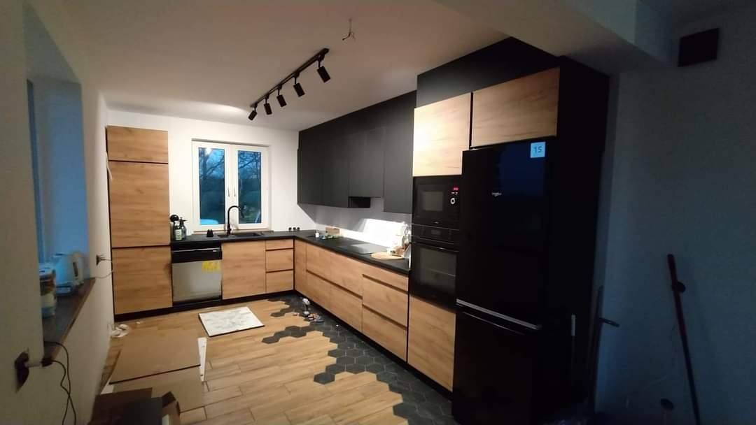 Nowoczesna kuchnia z drewnianymi frontami szafek dolnych i czarnymi szafkami górnymi, czarnym blatem i sprzętem AGD w zabudowie, widoczne fragmenty podłogi z drewnopodobnych paneli i heksagonalnych...