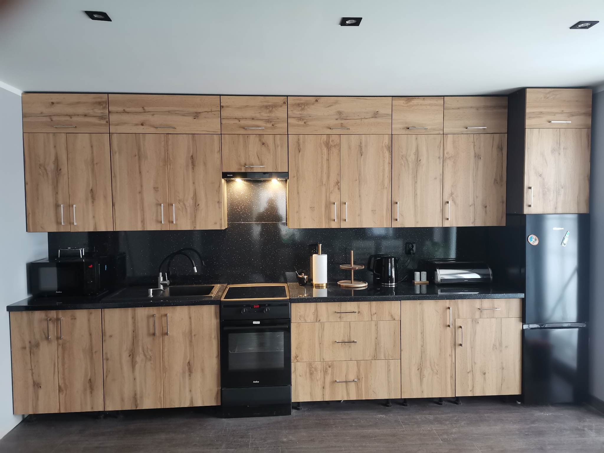 Kompletna zabudowa kuchenna z frontami imitującymi drewno, czarnym blatem i sprzętami AGD w nowoczesnym wnętrzu.