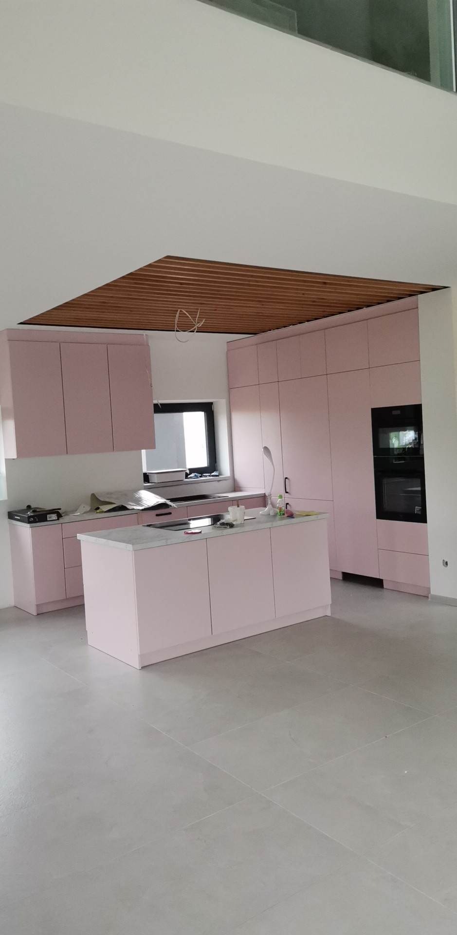 Różowa zabudowa kuchenna z wyspą i drewnianym sufitem w nowoczesnym wnętrzu, widoczne elementy montażowe.