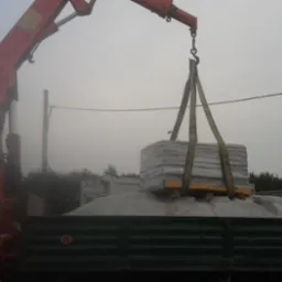 Ciężarówka z dźwigiem transportuje paletę kostki brukowej na tle zamglonego krajobrazu. Czerwony dźwig unosi ładunek nad wywrotką.