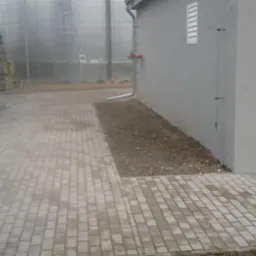 Ułożona kostka brukowa w przemysłowym otoczeniu, z widocznymi zbiornikami i fragmentem budynku. Szara kostka tworzy ścieżkę, kontrastując z ziemią obok.