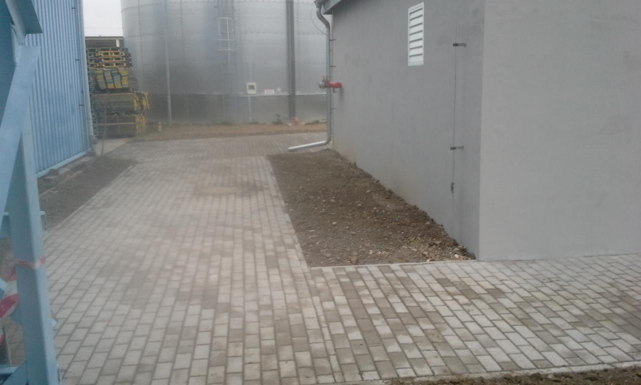 Ułożona kostka brukowa w przemysłowym otoczeniu, z widocznymi zbiornikami i fragmentem budynku. Szara kostka tworzy ścieżkę, kontrastując z ziemią obok.