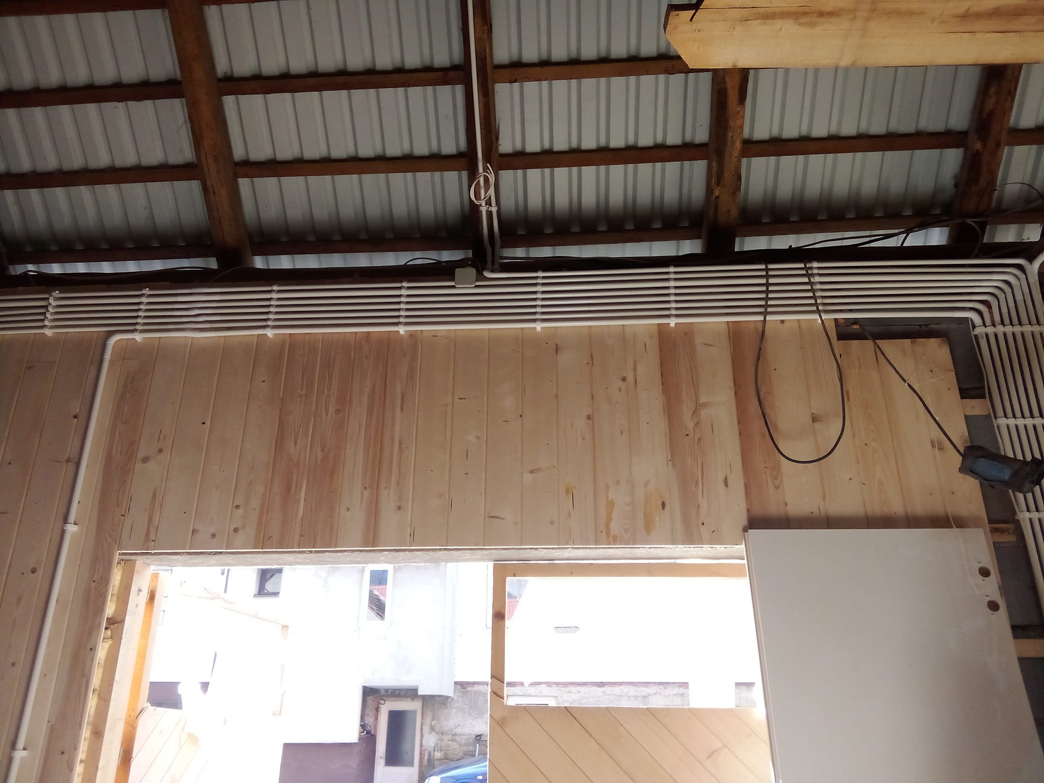 Białe rury instalacji elektrycznej ułożone równolegle pod blachą falistą dachu w drewnianym budynku w trakcie budowy, widok przez otwór okienny na zewnątrz.