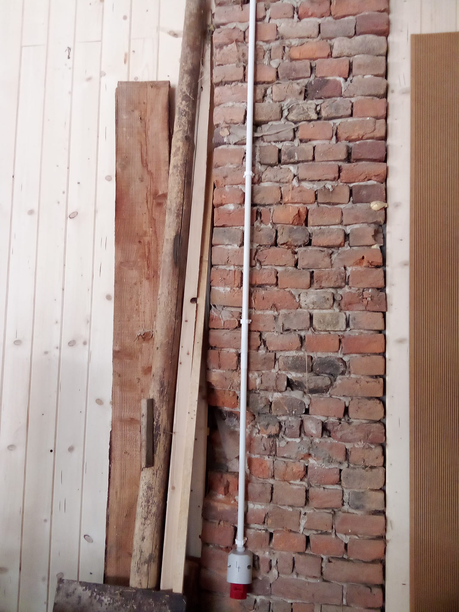 Wnętrze w trakcie remontu: fragment muru z cegły, pionowy kabel w białej osłonie, deski drewniane ułożone przy ścianie z boazerii.