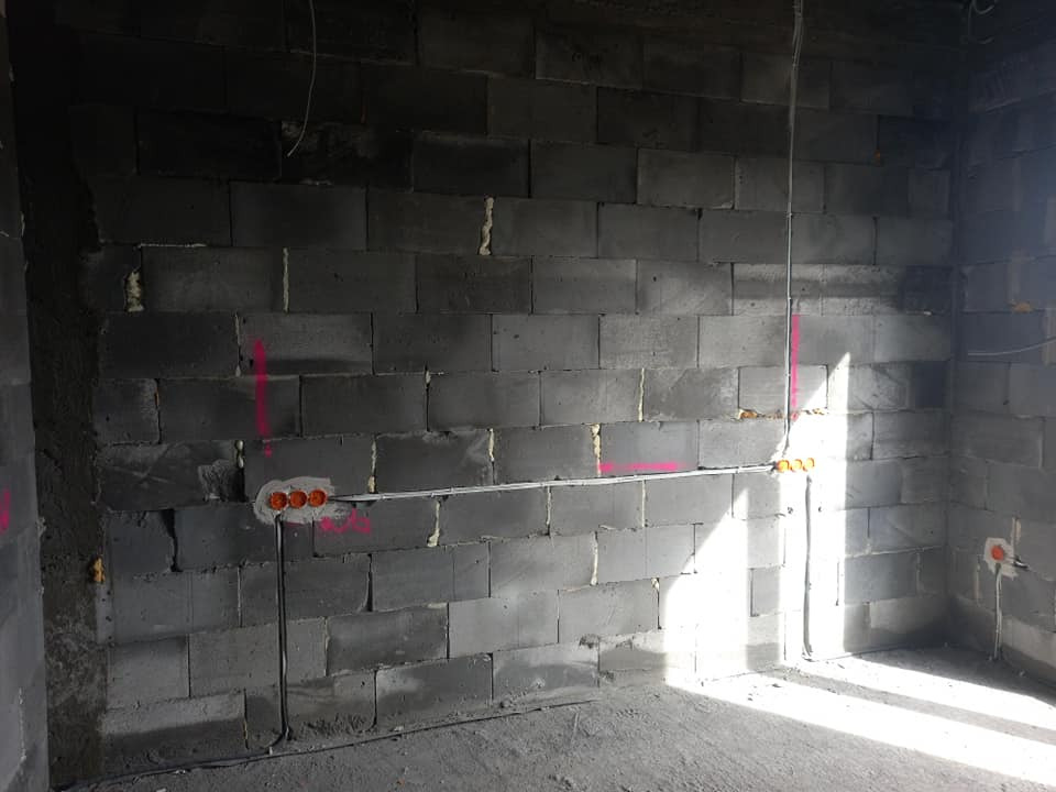 Surowa ściana z betonu komórkowego z widocznymi puszkami elektrycznymi, przewodami w peszlach i zaznaczeniami tras kabli różową farbą.