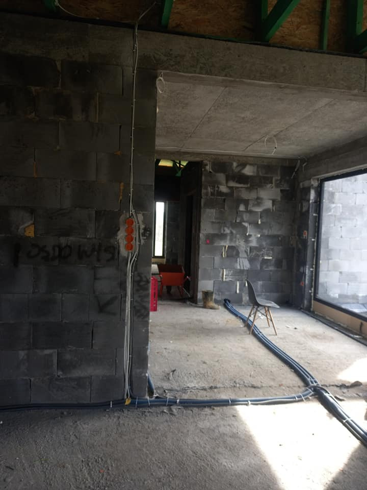 Surowy stan wnętrza z instalacją elektryczną: widoczne przewody w peszlach na podłodze i puszki elektryczne na ścianie z betonu komórkowego, czerwony wózek budowlany w tle.