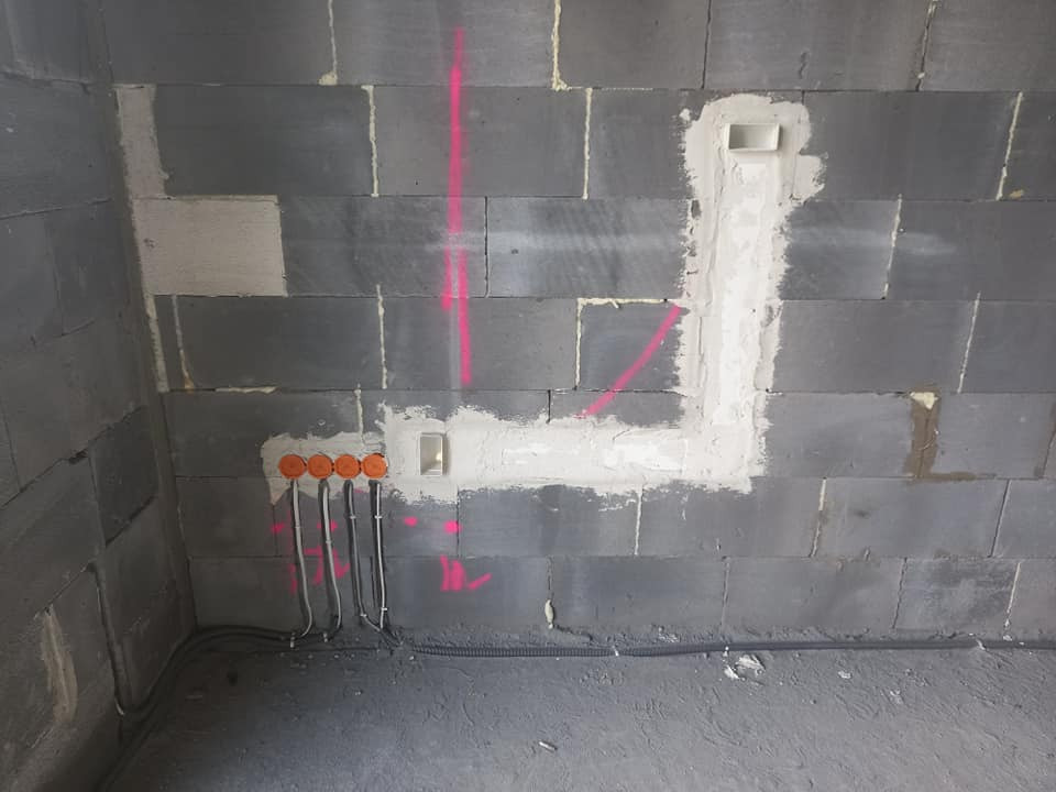 Surowa ściana z szarego betonu komórkowego z widocznymi przewodami elektrycznymi prowadzonymi w peszlach do pomarańczowych puszek instalacyjnych, zaznaczenia różowym markerem, wykuciem...