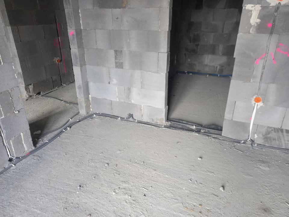 Surowy stan wnętrza w trakcie prac: szare ściany z betonu komórkowego z widocznymi instalacjami elektrycznymi w peszlach, różowe oznaczenia na ścianach, świeża wylewka podłogowa.