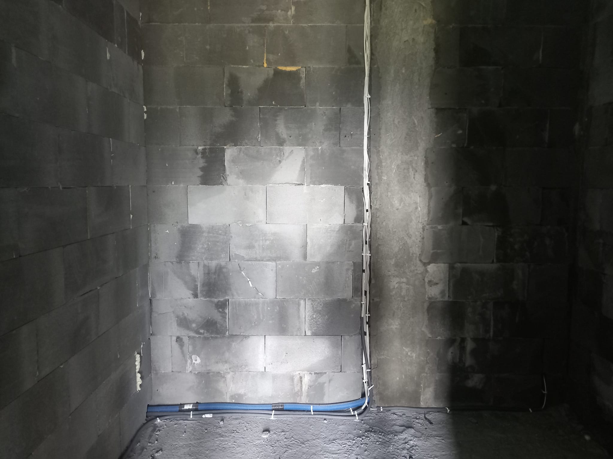 Surowa ściana z betonu komórkowego z widocznymi przewodami elektrycznymi i rurami w peszlu, zabezpieczonymi opaskami kablowymi przy podłodze.