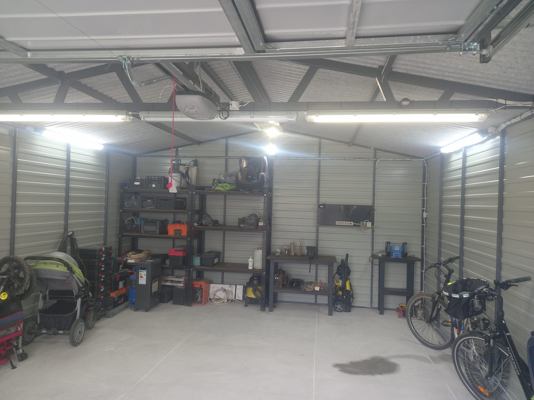 Wnętrze garażu z metalowymi ścianami, oświetlone lampami jarzeniowymi, z regałami pełnymi narzędzi i sprzętów, w tle widoczny automat do bramy garażowej.