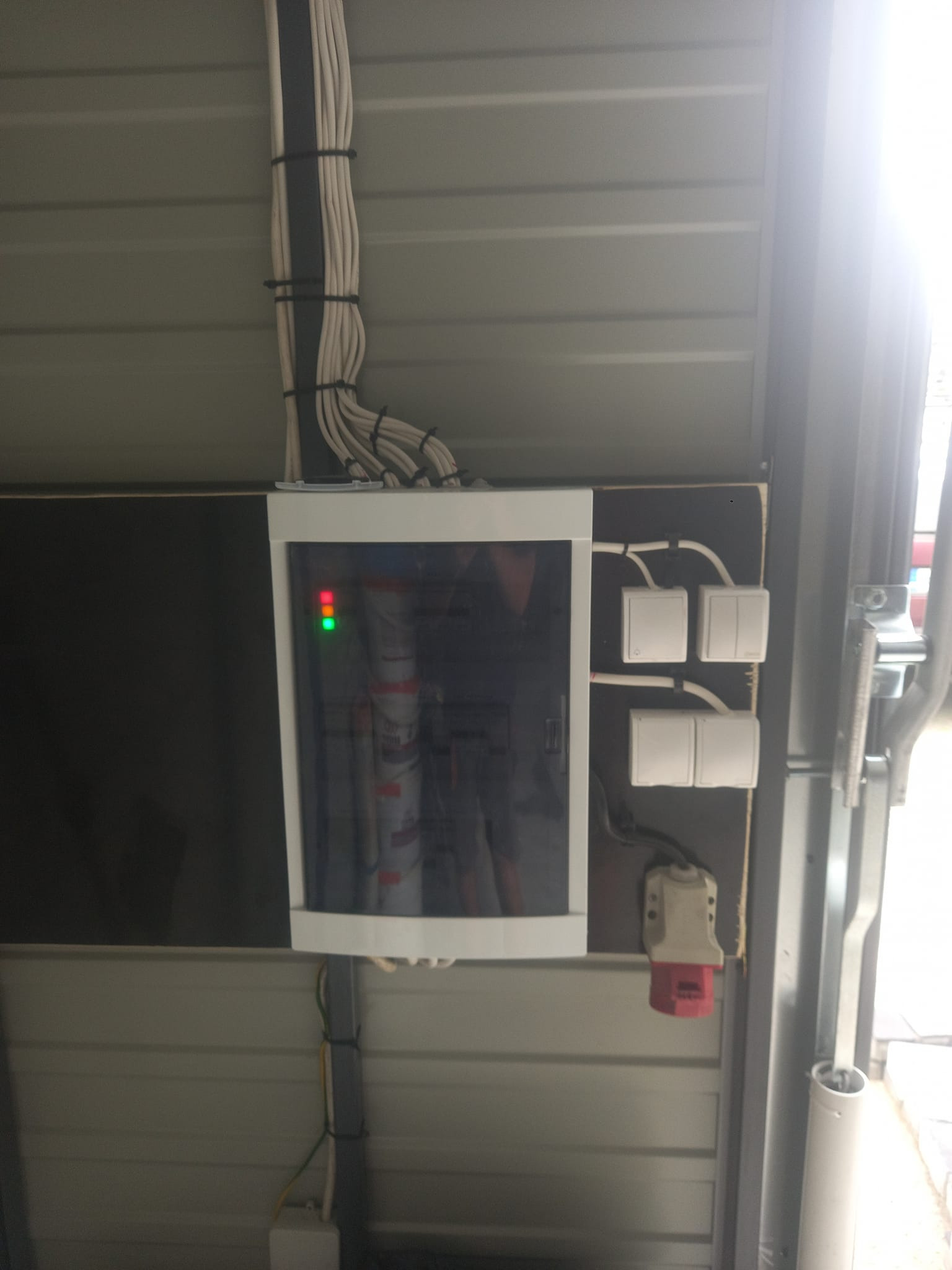 Rozdzielnica elektryczna z widocznymi przewodami, zabezpieczeniami i włącznikami zamontowana na ścianie z blachy trapezowej.