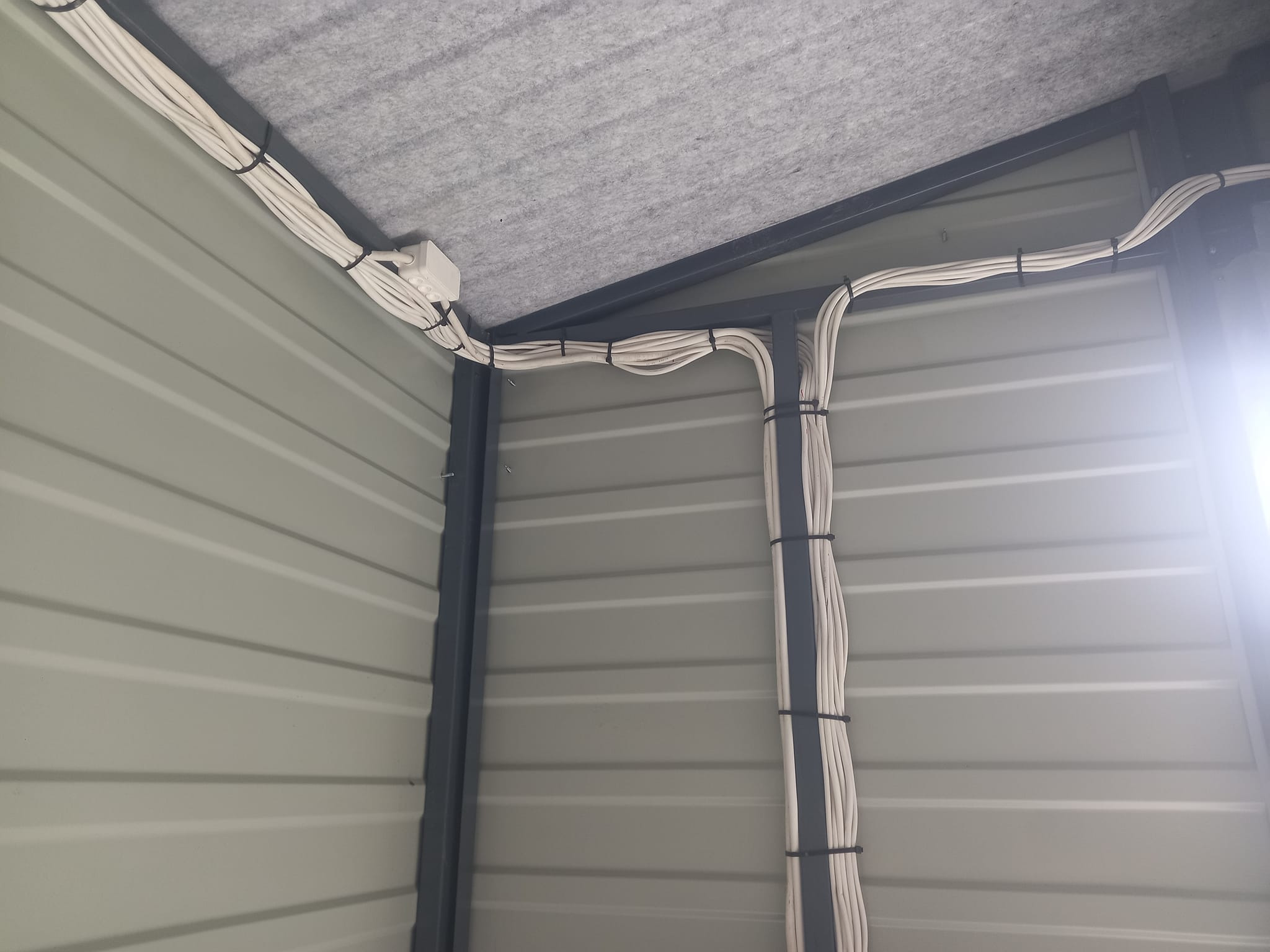 Wnętrze metalowej konstrukcji z widocznymi białymi kablami elektrycznymi, przymocowanymi czarnymi opaskami do metalowych ram, oraz szarą izolacją na suficie.