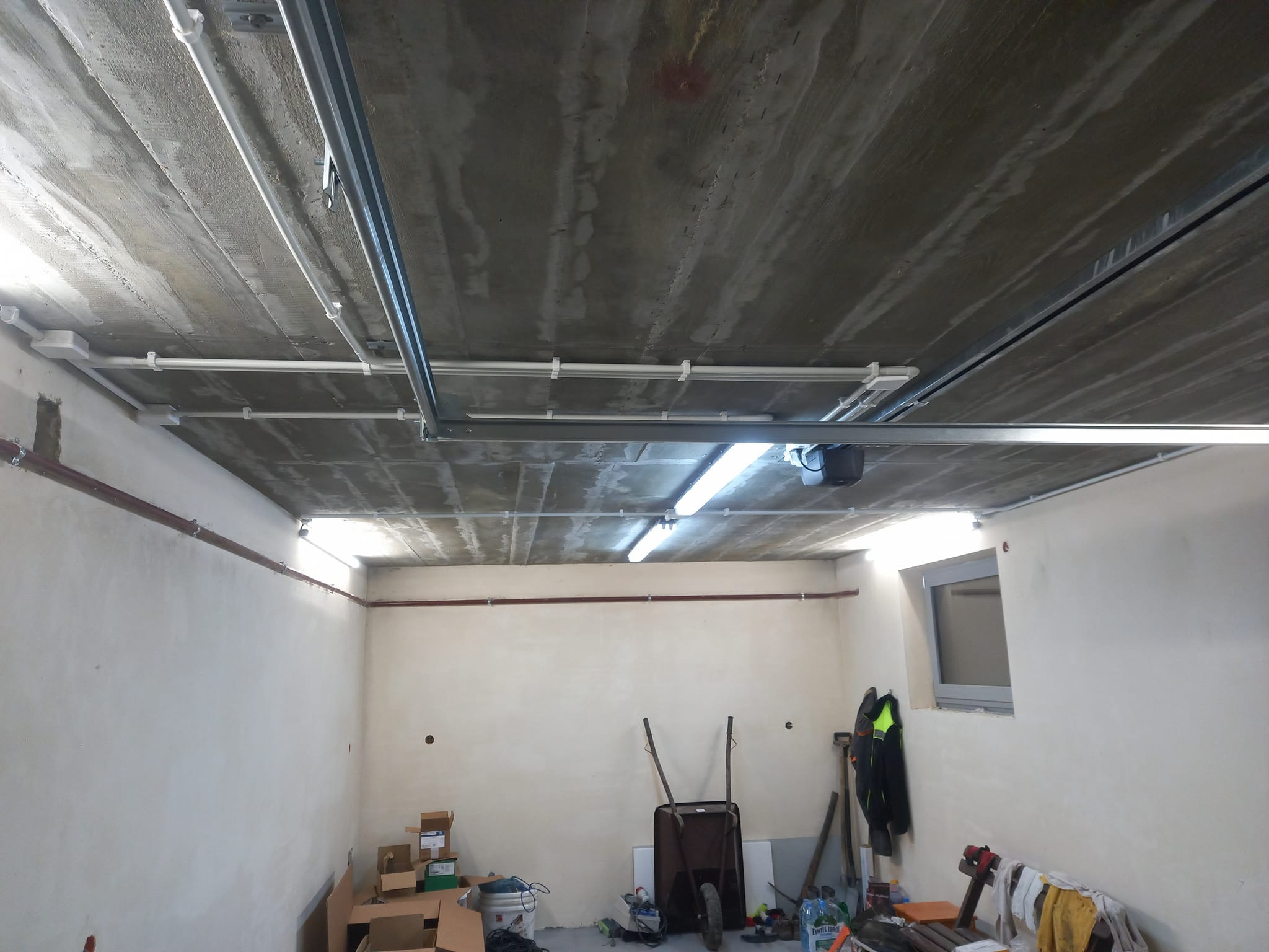 Surowy betonowy sufit garażu z widocznymi przewodami elektrycznymi w białych rurkach i metalowych korytkach, oświetlenie jarzeniowe, przygotowany do dalszych prac wykończeniowych.