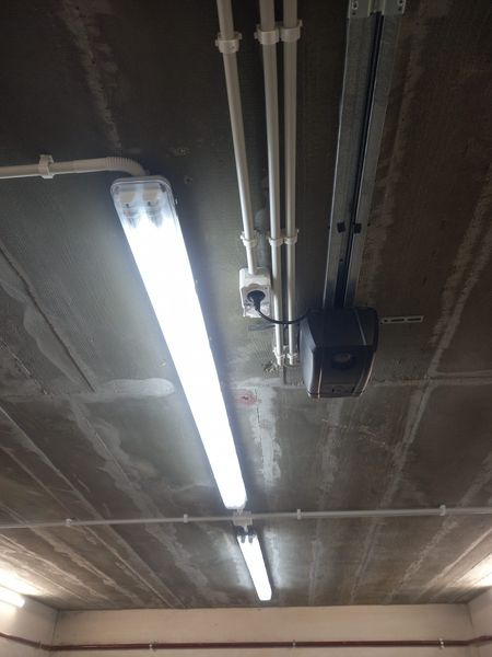 Widok sufitu z zamontowanym oświetleniem fluorescencyjnym, widoczne kable w białych rurkach PCV, puszka elektryczna z gniazdkiem i podłączonym przewodem, oraz mechanizm bramy garażowej na szynie...