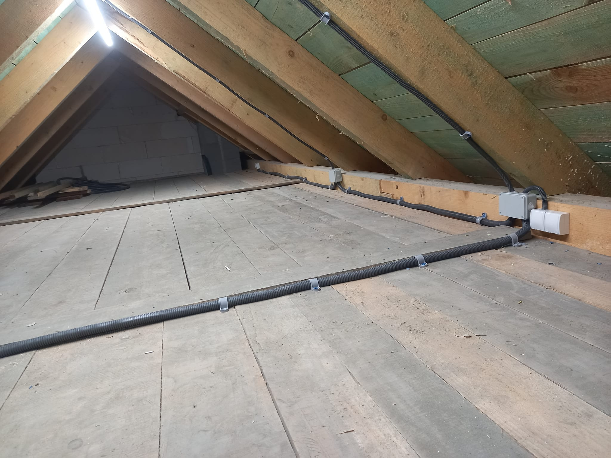Instalacja przewodów elektrycznych w karbowanej rurze ochronnej, przymocowanych klamrami do drewnianej konstrukcji poddasza, z widoczną puszką elektryczną i gniazdkami.