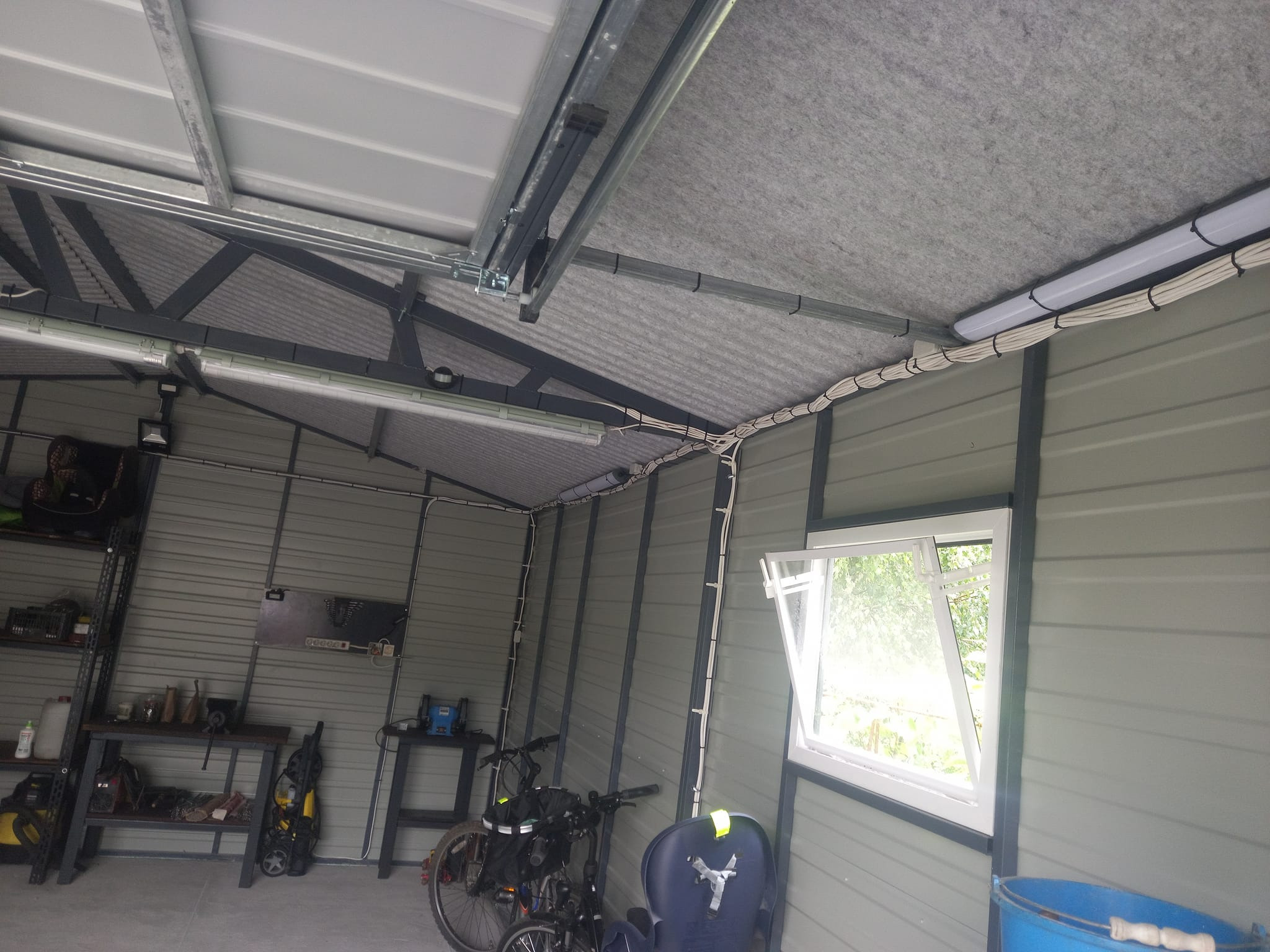 Wnętrze metalowego garażu z oknem, oświetleniem LED i instalacją elektryczną poprowadzoną po ścianach i suficie, widoczne rowery i wyposażenie warsztatowe.